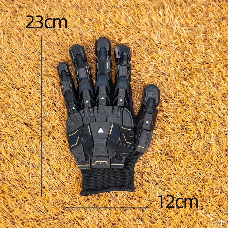 K&ouml;nnte beinhalten: Schwarz-goldener Handschuh mit futuristischem Design, 23 cm lang und 12 cm breit. Der Handschuh hat ein segmentiertes Aussehen mit einer schwarzen Manschette. Das Design beinhaltet geometrische Formen und goldene Details.
