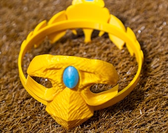 May include: A yellow crown with a blue gem at the center and top. The crown has a circular band and decorative elements. The crown is made of a textured material and sits on a brown surface.