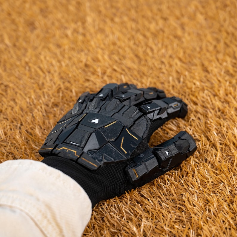 K&ouml;nnte beinhalten: Schwarzer Handschuh mit goldenen Akzenten und geometrischem Design. Der Handschuh besteht aus einem harten, segmentierten Material und wird &uuml;ber einer schwarzen Strickmanschette getragen. Der Hintergrund ist ein Feld aus trockenem, goldbraunem Gras.