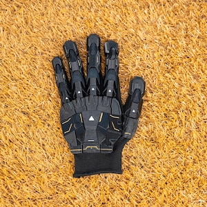 K&ouml;nnte beinhalten: Ein schwarzer und goldener Roboterhandschuh mit beweglichen Fingern. Der Handschuh hat ein strukturiertes, segmentiertes Design mit goldenen Akzenten. Der Handgelenkbereich ist ein schwarzes, geripptes Gewebe. Der Handschuh liegt auf Kunstgras.