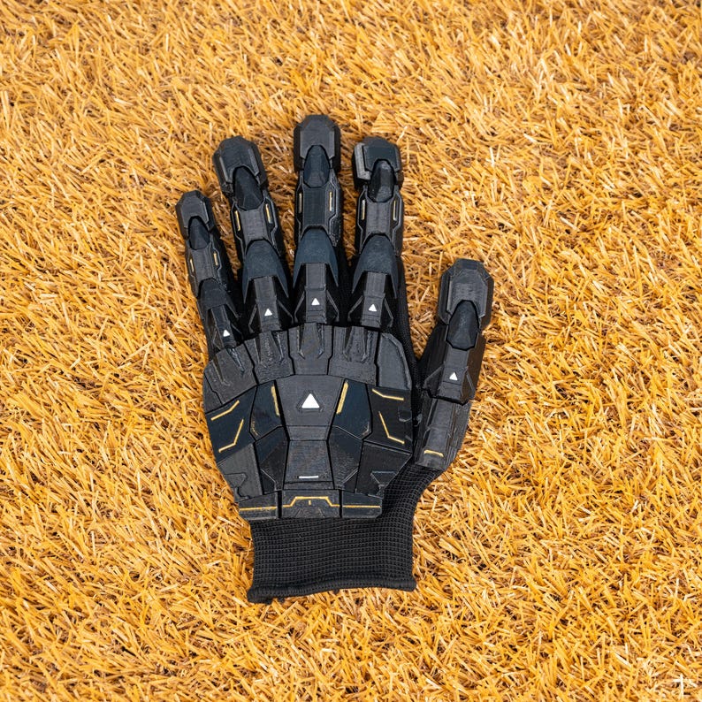 K&ouml;nnte beinhalten: Ein schwarzer und goldener Roboterhandschuh mit beweglichen Fingern. Der Handschuh hat ein strukturiertes, segmentiertes Design mit goldenen Akzenten. Der Handgelenkbereich ist ein schwarzes, geripptes Gewebe. Der Handschuh liegt auf Kunstgras.