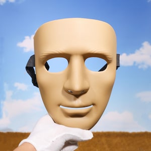 May include: A tan-coloured, full-face mask with eye and mouth openings. The mask has a realistic human face design, featuring a nose, and a smiling mouth. A black strap is attached on each side. The mask is held by a gloved hand.