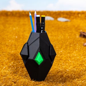 Op de afbeelding: Een zwarte geometrische pennenhouder met een groene ruitvormige accent. De houder bevat een blauw potlood, een paars potlood en twee zwarte markers met witte en gele accenten. De pennenhouder staat op geel gras.