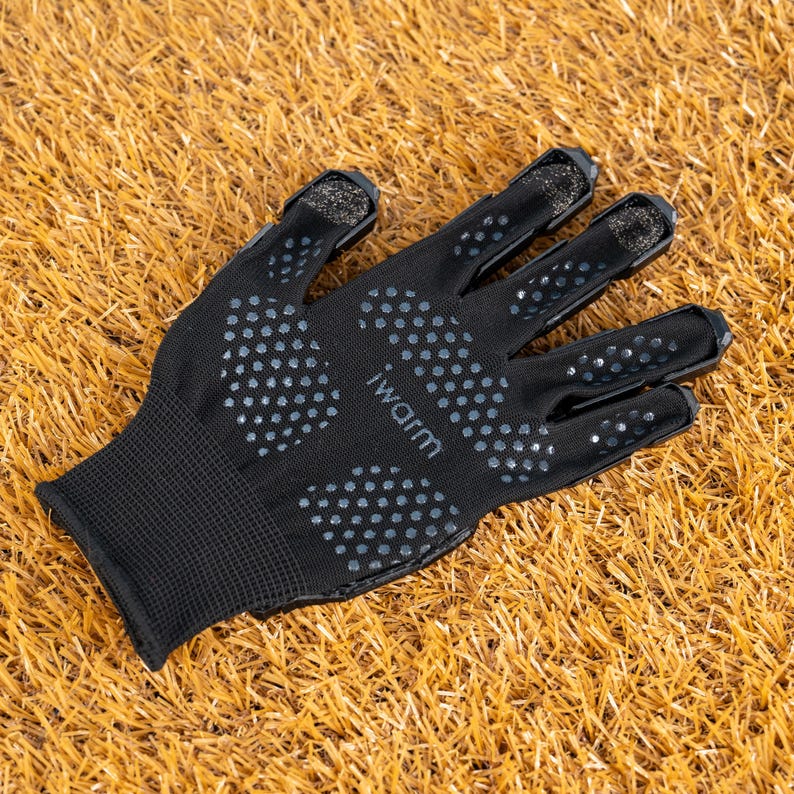 K&ouml;nnte beinhalten: Schwarzer Handschuh mit grauem Punktmuster und dem Text "iWarm" am Handgelenk. Der Handschuh hat verst&auml;rkte Fingerspitzen und eine strukturierte Handfl&auml;che f&uuml;r besseren Halt. Der Handschuh liegt auf braunem Gras.