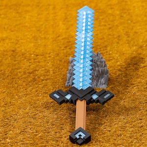 Puede incluir: Una espada de juguete pixelada con una hoja azul claro y un mango marrón. La empuñadura es negra con detalles en azul claro. La espada está sobre una superficie texturizada de color marrón dorado.
