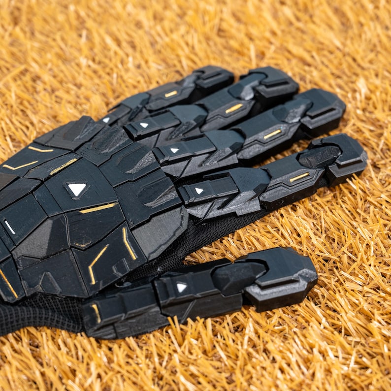 K&ouml;nnte beinhalten: Schwarzer Roboterhandschuh mit geometrischen Paneelen und goldenen Akzenten. Der Handschuh liegt auf trockenem, goldbraunem Gras. Das Design weist eckige Formen und kleine wei&szlig;e Dreieckdetails auf.