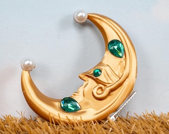 May include: Gold-colored crescent moon hair clip with a face design, adorned with green gemstone accents and two faux pearls. The hair accessory is secured with a silver metal clip, suitable for various hairstyles.