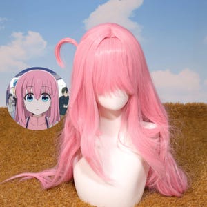 Puede incluir: Una peluca rosa larga con flequillo y una pequeña cola de caballo, exhibida en una cabeza de maniquí. La peluca está peinada con capas largas y fluidas. Una imagen de un personaje de anime está en el fondo.