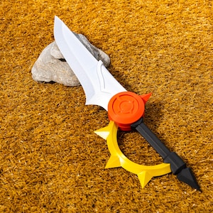 May include: A toy sword with a white blade and a red and yellow hilt. The hilt features a circular red guard with a stylized design and a yellow, star-shaped guard. The sword is resting on a bed of golden-brown material.