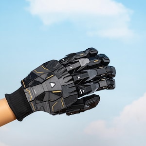 K&ouml;nnte beinhalten: Ein schwarzer und grauer Roboterhandschuh mit geometrischen Paneelen und goldenen Akzenten. Der Handschuh wird &uuml;ber einer schwarzen Strickmanschette getragen. Der Hintergrund ist ein blauer Himmel mit Wolken.