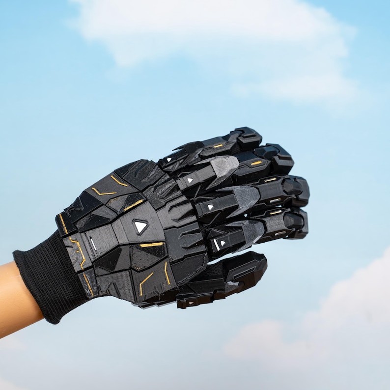K&ouml;nnte beinhalten: Ein schwarzer und grauer Roboterhandschuh mit geometrischen Paneelen und goldenen Akzenten. Der Handschuh wird &uuml;ber einer schwarzen Strickmanschette getragen. Der Hintergrund ist ein blauer Himmel mit Wolken.
