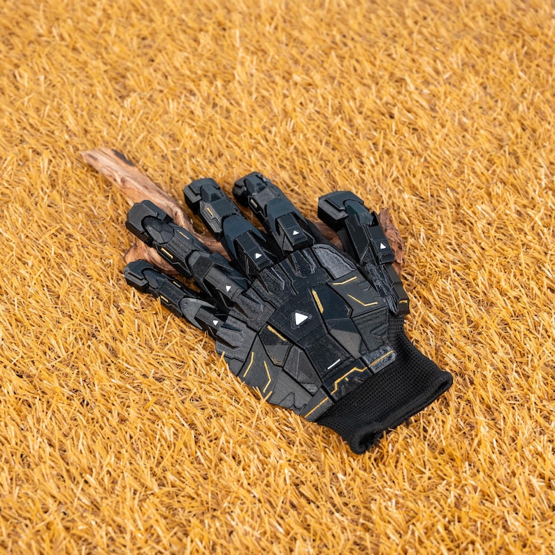 K&ouml;nnte beinhalten: Ein schwarzer und goldener Roboterhandschuh mit strukturiertem Design. Der Handschuh hat geometrische Formen und eine schwarze Handgelenkmanschette. Er liegt auf einem Bett aus trockenem, goldbraunem Gras.