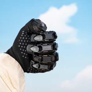 K&ouml;nnte beinhalten: Schwarzer Handschuh mit strukturierter Handfl&auml;che und einer Reihe von beweglichen, schwarzen Panzerplatten, die die Kn&ouml;chel und Finger bedecken. Der Handschuh wird zu einer Faust geballt vor blauem Himmel mit Wolken gehalten.