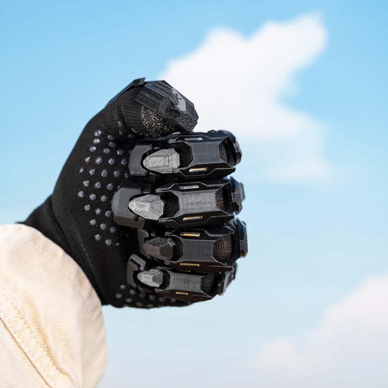 K&ouml;nnte beinhalten: Schwarzer Handschuh mit strukturierter Handfl&auml;che und einer Reihe von beweglichen, schwarzen Panzerplatten, die die Kn&ouml;chel und Finger bedecken. Der Handschuh wird zu einer Faust geballt vor blauem Himmel mit Wolken gehalten.