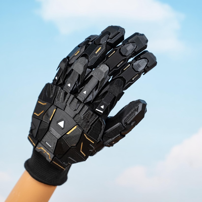 K&ouml;nnte beinhalten: Schwarzer Roboterhandschuh mit geometrischen Formen und goldenen Akzenten. Der Handschuh wird auf einer Hand vor blauem Himmel getragen. Das Design weist gegliederte Segmente und eine strukturierte Oberfl&auml;che auf, was auf ein futuristisches oder Cosplay-Thema hindeutet.