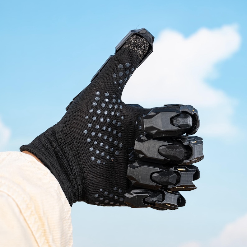 K&ouml;nnte beinhalten: Schwarzer Handschuh mit strukturierter Oberfl&auml;che und silbernen Punkten, mit starren schwarzen Schutzelementen an Kn&ouml;cheln und Fingern. Der Daumen ist als Zeichen der Zustimmung vor blauem Himmel ausgestreckt.