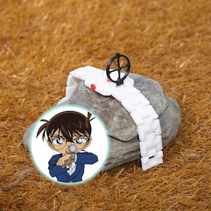 May include: A white wristwatch with a black dial and a red button is displayed on a rock. The watch has a white band. A cartoon character is in the lower left corner. The background is brown.