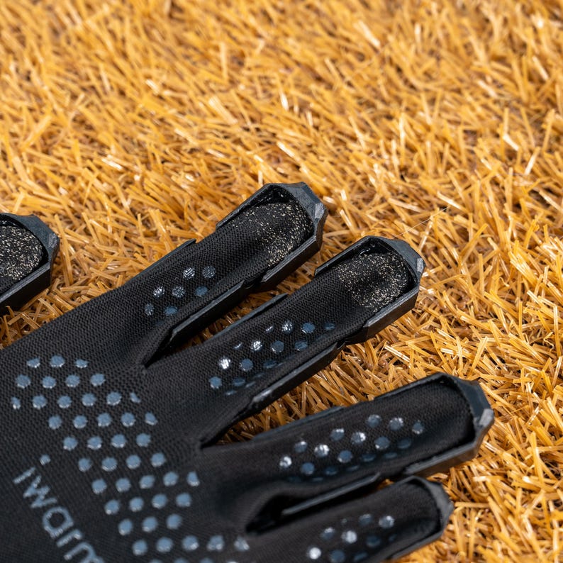 K&ouml;nnte beinhalten: Nahaufnahme eines schwarzen Handschuhs mit einem gepunkteten Muster und dem Text "iWarm". Der Handschuh hat Mesh-Details an den Fingern und liegt vor einem Hintergrund aus trockenem, goldbraunem Gras.