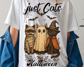 Könnte beinhalten: Weißes T-Shirt mit einem Halloween-Motiv. Das Design zeigt drei Cartoon-Katzen in Kostümen: eine Mumie, ein Geist und eine Hexe. Der Text lautet "just Cats Halloween". Fledermäuse und Spinnweben sind ebenfalls enthalten.