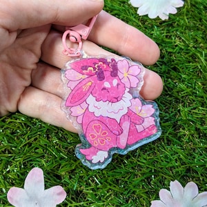 Op de afbeelding: Een roze acryl sleutelhanger met een cartoon konijn met bloemmotieven. Het konijn heeft een witte, pluizige borst en is omringd door roze kersenbloesems. De sleutelhanger heeft een roze sluiting en wordt vastgehouden tegen een achtergrond van groen gras en bloemblaadjes.