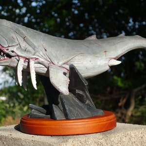 May include: A gray and white whale sculpture with a menacing open mouth and sharp teeth. The sculpture is mounted on a black rock base and a wooden platform.