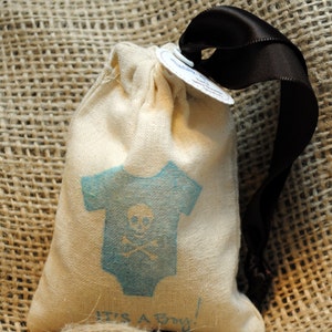 May include: A small, white fabric bag with a blue and white baby onesie design. The onesie has a skull and crossbones design. The bag has a black ribbon tied around it and a tag that says "It's a Boy!"