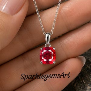 May include: A silver necklace featuring a cushion-cut vibrant red gemstone pendant held by four prongs on a delicate chain. The text SparklegemsArt is visible near the pendant. The design is simple, focusing on the bright red stone and metal.