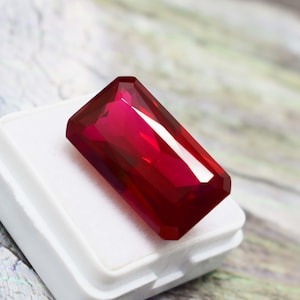 May include: A vibrant, deep red gemstone with a rectangular emerald cut, featuring multiple facets that reflect light. The stone is positioned on a small, white, square display base, showcasing its intense colour and clear, polished surface.