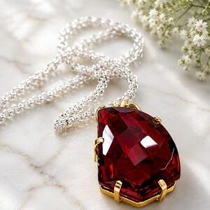 May include: A necklace featuring a large, faceted, pear-shaped deep red gemstone set in a gold-toned metal prong setting, attached to a silver-toned chain. The text SparklegemsArt is printed in black at the bottom of the image.