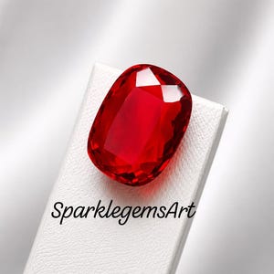 May include: A vibrant, cushion-cut red gemstone with multifaceted surfaces that reflect light. The gemstone is positioned on a white, textured display stand featuring the black text SparklegemsArt printed clearly on the lower portion of the surface.