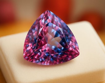 May include: A triangular-cut gemstone with facets, displaying a blend of pink and blue hues. The gem sits on a white, rectangular surface, showcasing its polished appearance. The gem's color transitions from pink to blue.