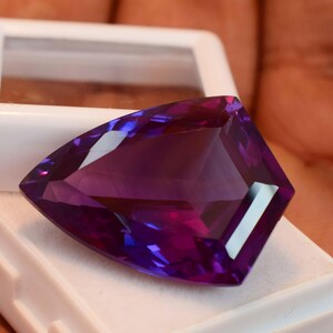 May include: A faceted, shield-shaped purple gemstone with a brilliant cut, displaying deep violet hues and sharp geometric edges. The stone is positioned resting on a white plastic display case with a clear lid, highlighting its translucent clarity.