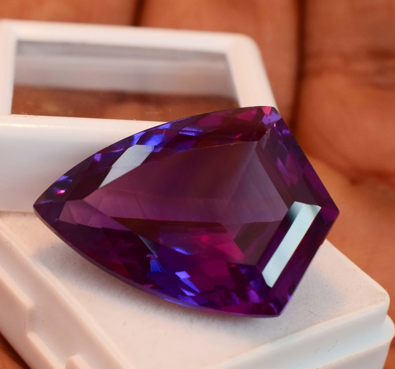 May include: A faceted, shield-shaped purple gemstone with a brilliant cut, displaying deep violet hues and sharp geometric edges. The stone is positioned resting on a white plastic display case with a clear lid, highlighting its translucent clarity.
