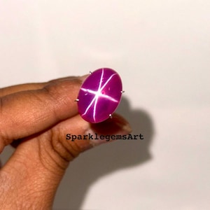 May include: A vibrant, oval-shaped, star ruby gemstone with a six-ray star pattern. The gemstone is a deep magenta color with a bright white star. The gemstone is set in a silver metal setting. The text "SparklegemsArt" is visible.