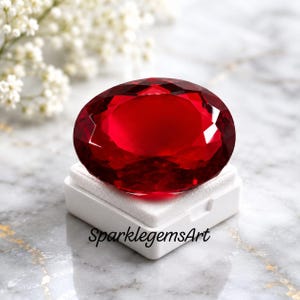 May include: A large, oval-cut, deep red gemstone with multifaceted surfaces that reflect light. The gemstone rests on a white square display base with the text SparklegemsArt printed in black on the front side of the white base.