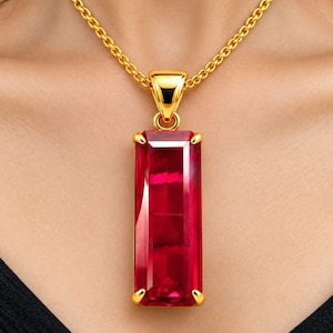 Può includere: Una collana a catena dorata con un grande ciondolo rettangolare in pietra preziosa rossa sfaccettata, tenuto da quattro griffe dorate. Il ciondolo è sospeso a una contromaglia dorata. Il testo SparklegemsArt è visibile in basso.