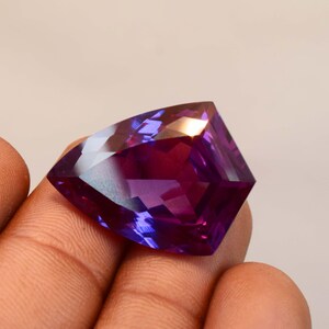 May include: A faceted, shield-shaped gemstone displaying a vibrant colour gradient that shifts from deep purple to magenta. The stone features a polished, multi-faceted surface that reflects light, creating distinct geometric patterns across its face.