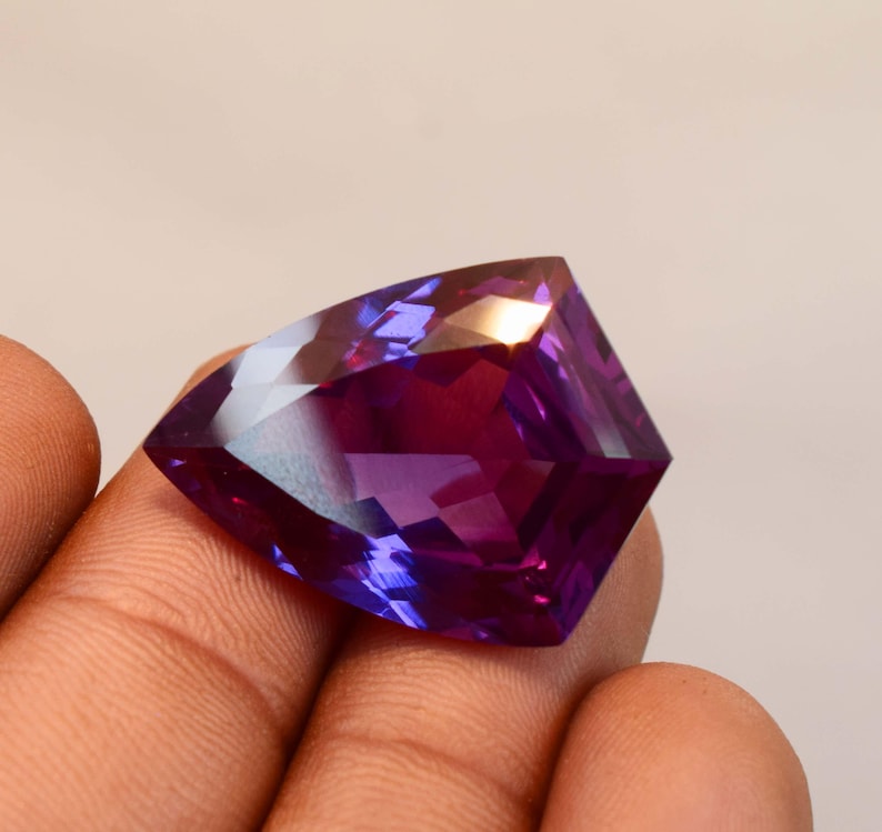 May include: A faceted, shield-shaped gemstone displaying a vibrant colour gradient that shifts from deep purple to magenta. The stone features a polished, multi-faceted surface that reflects light, creating distinct geometric patterns across its face.