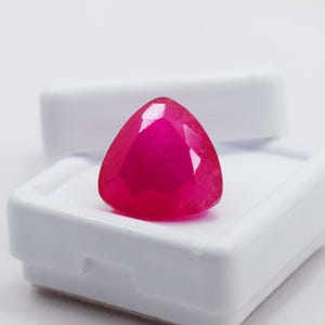 May include: A vibrant, deep pink gemstone cut into a triangular shape with multiple facets that reflect light. The stone is positioned on a white, rectangular display box with a hinged lid, showcasing its saturated color and geometric form.