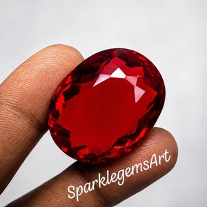 May include: A large, oval-cut, vibrant red gemstone with multifaceted surfaces that reflect light. The stone features a deep crimson hue and is held in place. The text SparklegemsArt is printed in white script across the lower portion of the gem.