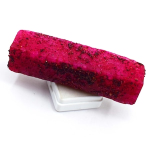 Può includere: Un cristallo di pietra preziosa grezza di forma rettangolare, color magenta vibrante, con una superficie cristallina testurizzata che presenta inclusioni scure e punteggiate lungo tutta la sua lunghezza, poggiato su una piccola base quadrata in plastica bianca.