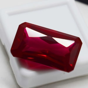 May include: A vibrant, deep red gemstone cut into a rectangular shape with faceted edges. The stone displays high clarity and a polished surface, reflecting light across its top facets. It is positioned resting on a small, square white display case.