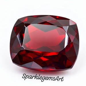 May include: A deep red, cushion-cut gemstone with multiple triangular and trapezoidal facets that reflect light. The stone has a saturated crimson hue and a polished, glassy surface. The text SparklegemsArt is printed in black below the gem.