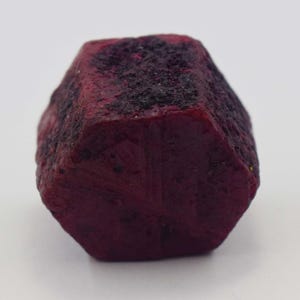 May include: A raw, hexagonal ruby crystal with a deep, saturated red color. The surface of the gemstone displays a natural, unpolished texture with dark, rough inclusions and geometric facets that define its distinct six-sided crystalline shape.