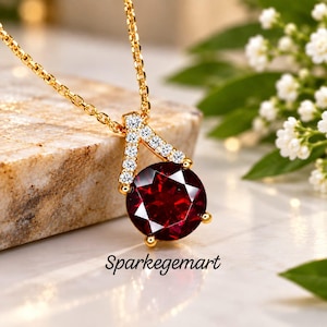 May include: A gold necklace featuring a round, deep red gemstone pendant suspended from a triangular bail accented with small clear stones. The text Sparkegemart is printed below the pendant. The chain is a delicate gold link style.