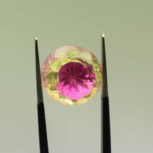 May include: A round, faceted gemstone held by tweezers. The gem has a colour gradient, with a deep pink centre and a yellow-green outer edge. The tweezers are black.