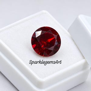 Puede incluir: Una piedra preciosa redonda y facetada de color rojo intenso presentada en una caja cuadrada blanca. La piedra tiene un corte brillante con múltiples superficies reflectantes. El texto SparklegemsArt está impreso en negro sobre la espuma.
