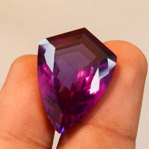 May include: A faceted shield-shaped gemstone with a deep purple and magenta colour gradient. The stone features a polished surface with multiple geometric facets that reflect light, creating a vibrant and translucent appearance throughout the crystal.