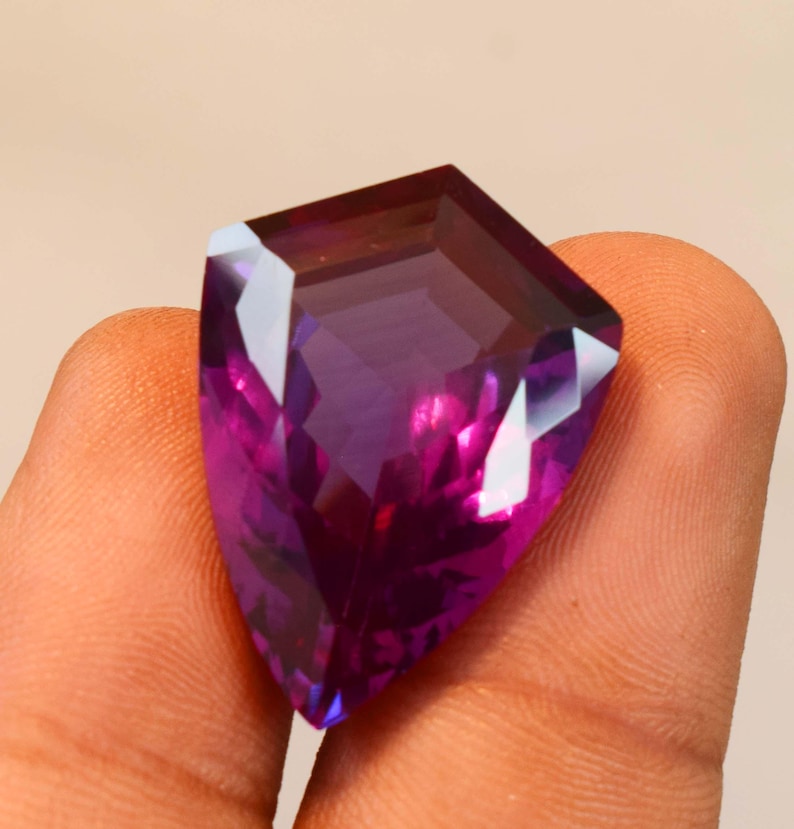 May include: A faceted shield-shaped gemstone with a deep purple and magenta colour gradient. The stone features a polished surface with multiple geometric facets that reflect light, creating a vibrant and translucent appearance throughout the crystal.