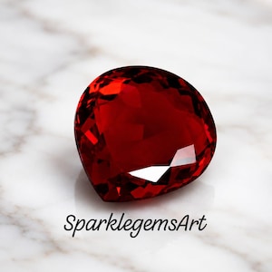 May include: A deep red, heart-shaped gemstone with a faceted surface that reflects light. The stone is positioned above the black text SparklegemsArt, which is written in a cursive font. The gem displays a rich, saturated crimson colour.